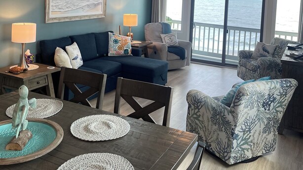 A living room with a view of the ocean, a ceiling fan, and a dining table with placemats. - Oceanfront 2Br 2Ba Condo with elevator and amazing views! (Surfside Beach)