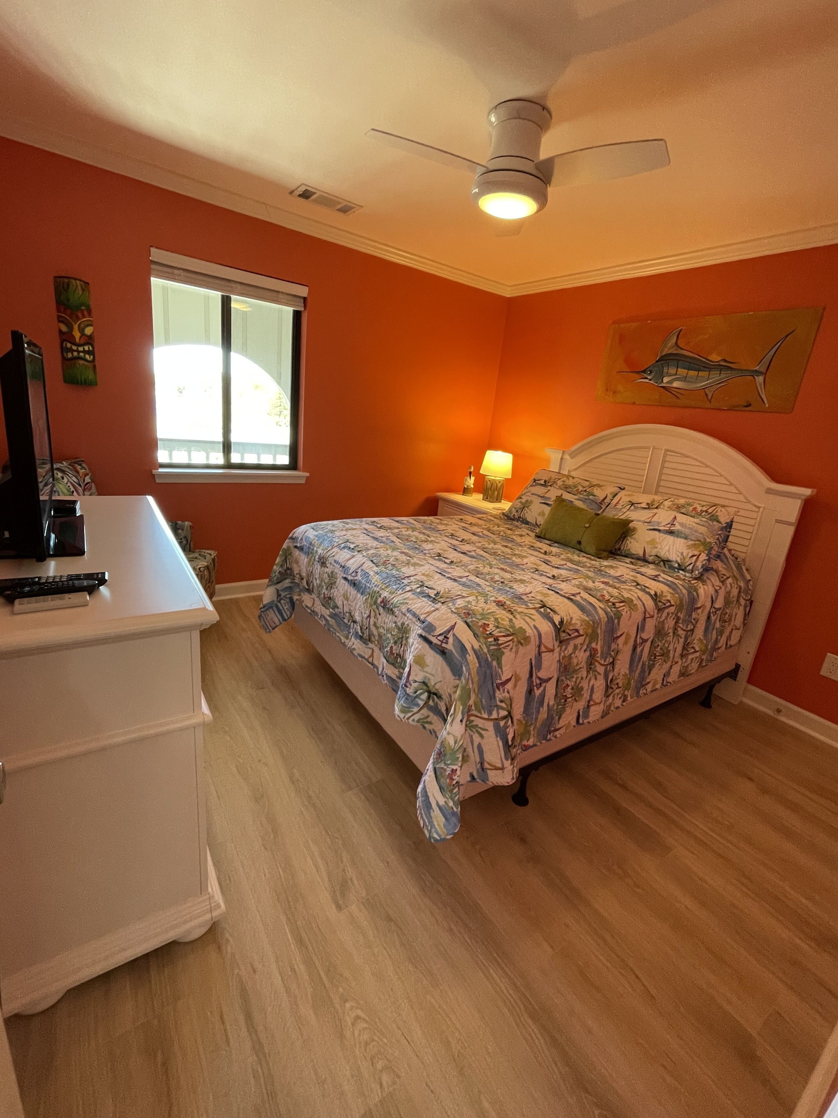 Un dormitorio con cama, escritorio, ventilador de techo, ventana y un cuadro en la pared.
