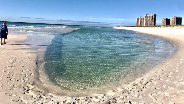 Ein Strand mit klarem Wasser, Fußabdrücken und hohen Gebäuden im Hintergrund.