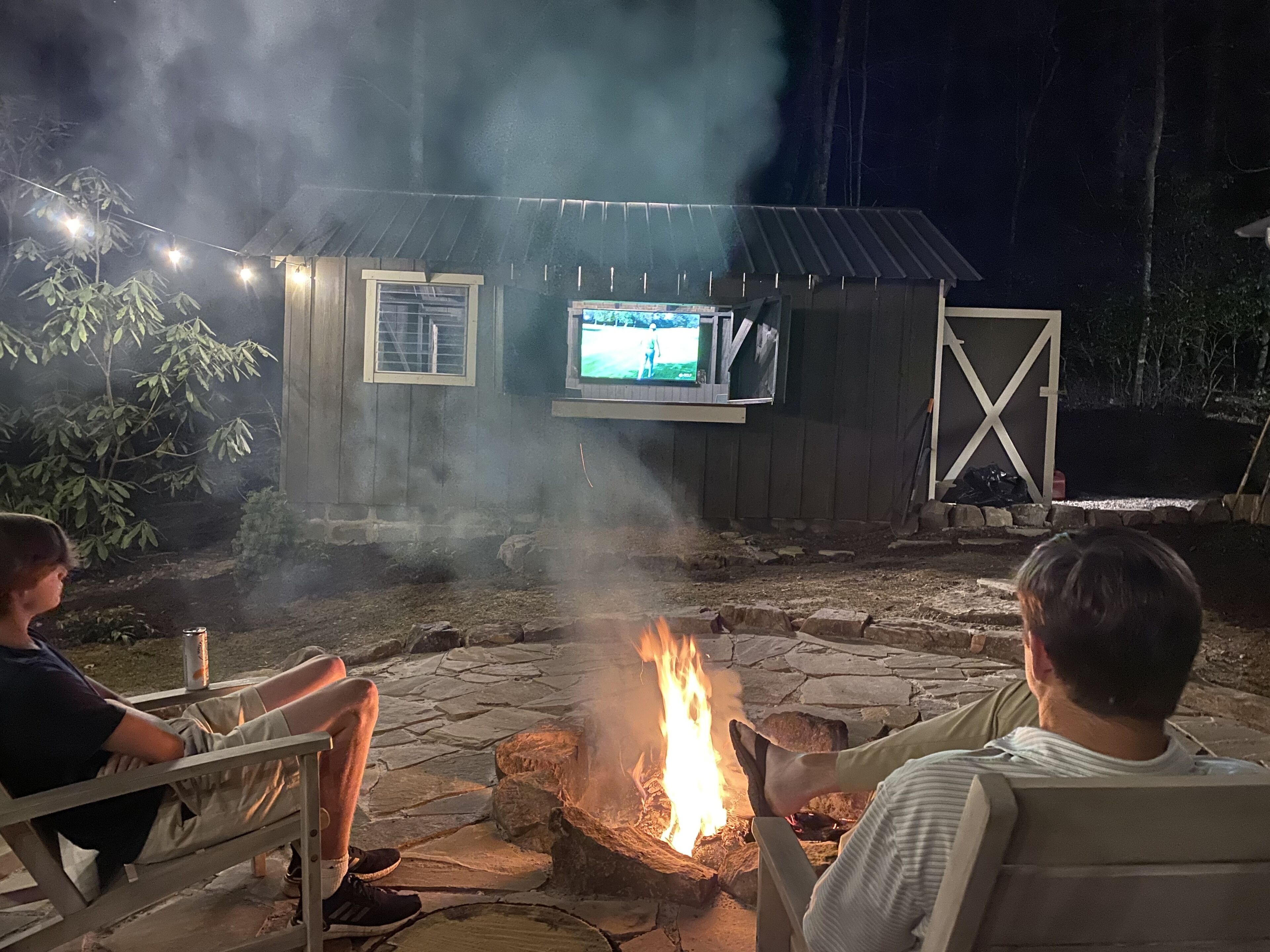 A stone fire pit with a television screen mounted on a wooden structure, surrounded by stone steps and a metal gate.