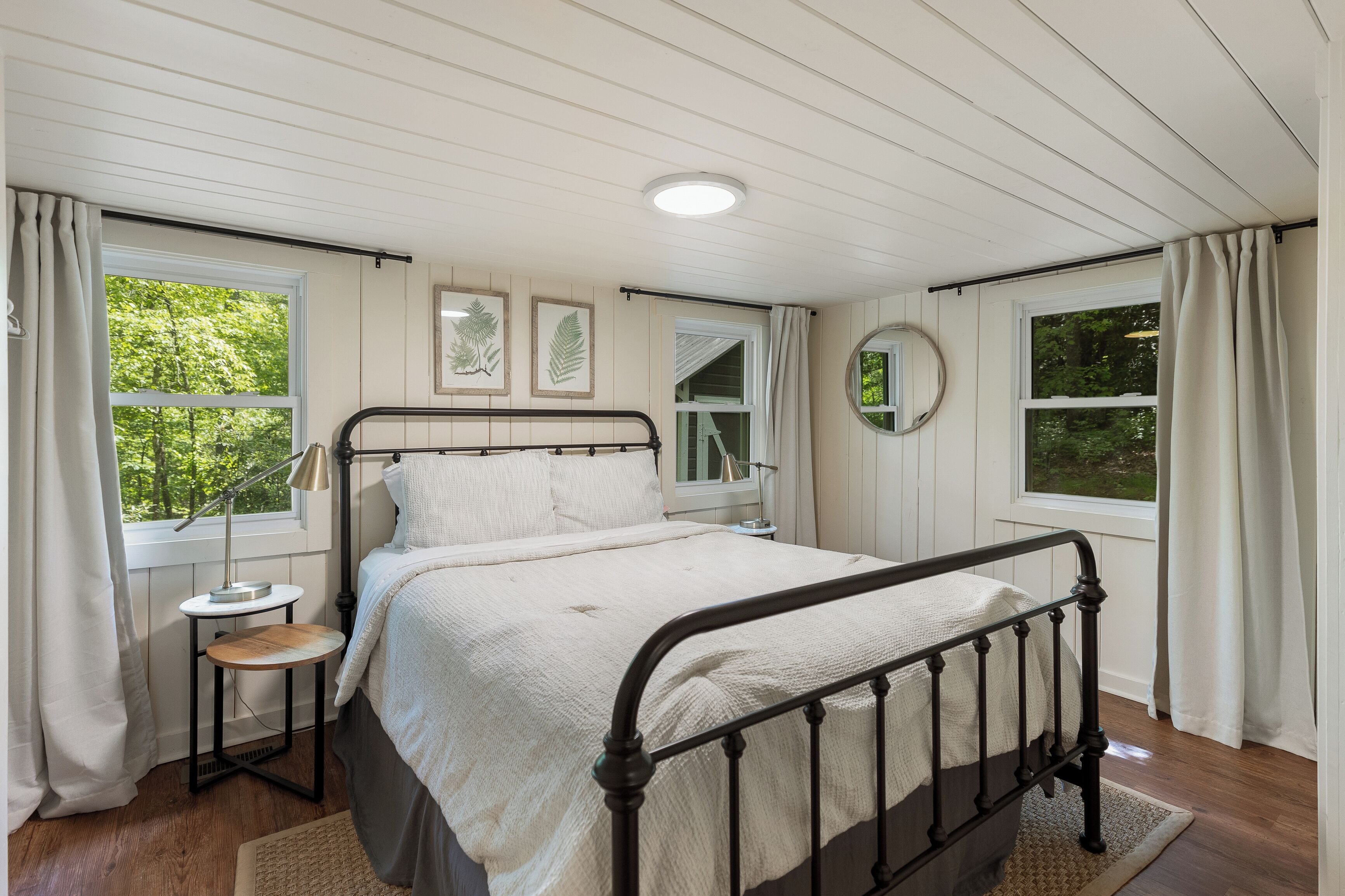Une chambre à coucher avec un lit, des tables de chevet, un grand miroir rond et de grandes fenêtres avec des rideaux.