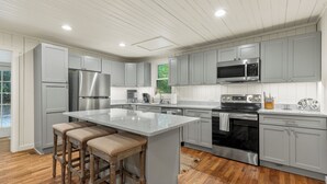 A modern kitchen with grey cabinets, stainless steel appliances, a large island with bar stools, and wooden flooring.