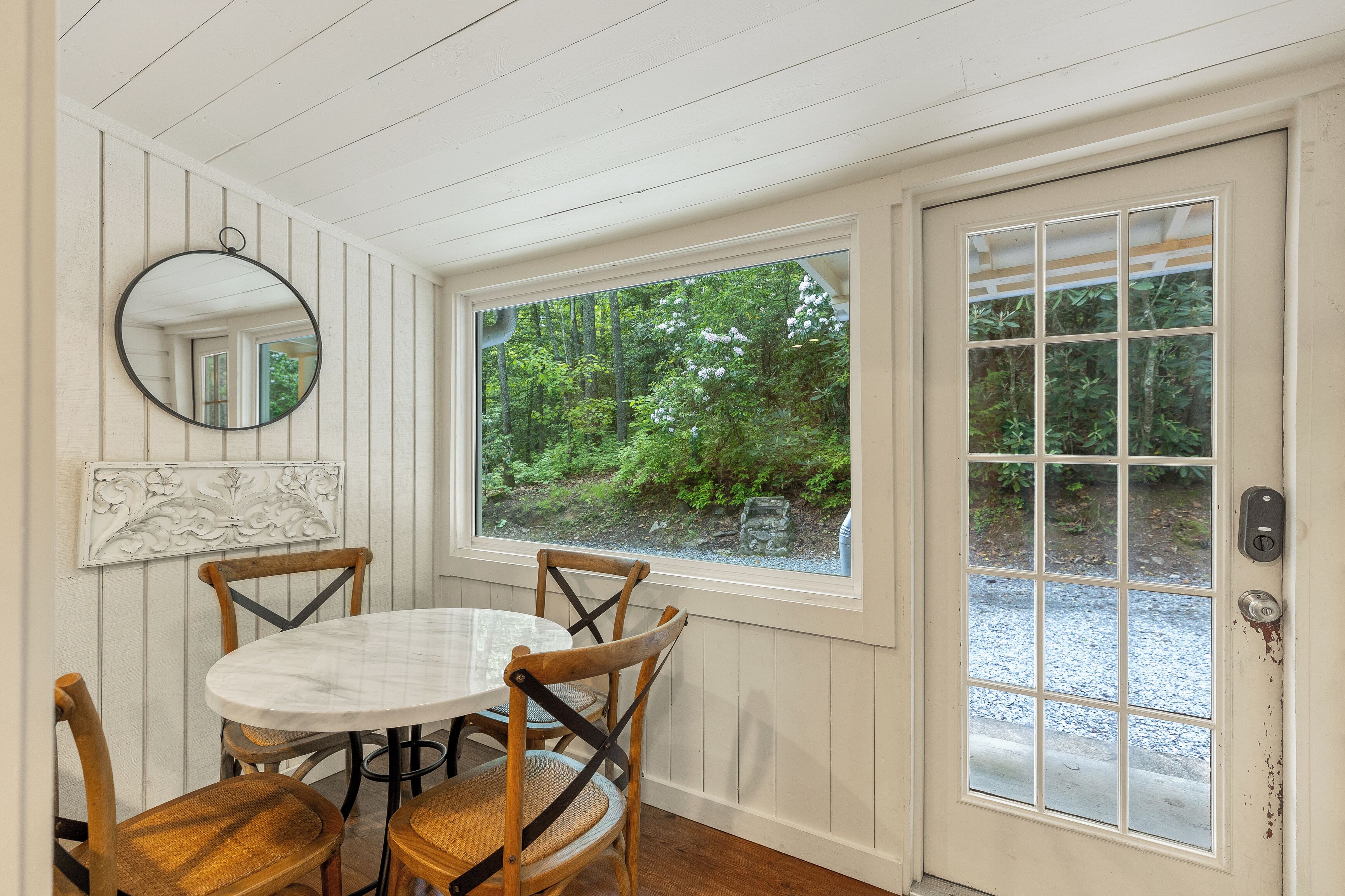 A dining area with a round table, four wooden chairs, a white marble tablecloth, a round mirror, and a window with a view of greenery.