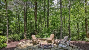 A stone fire pit with Adirondack chairs in a forest setting.
