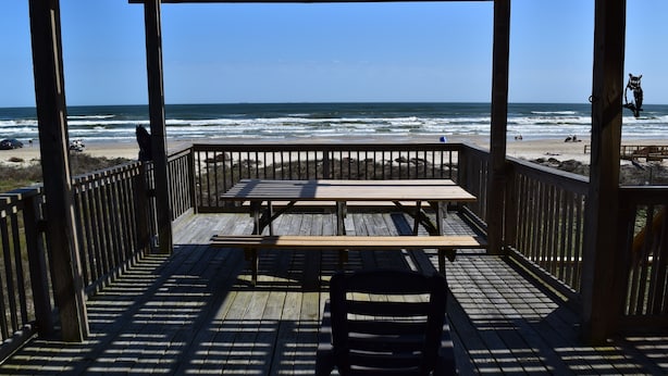 A wooden deck with a picnic table and chairs, overlooking a beach with waves and people in the distance. - Amazing 5-bedroom beachfront home waiting for your getaway, sleeps 16 (Surfside Beach)