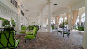 Un patio interior espacioso con sillas y mesas verdes, columnas blancas y suelo de ladrillo.