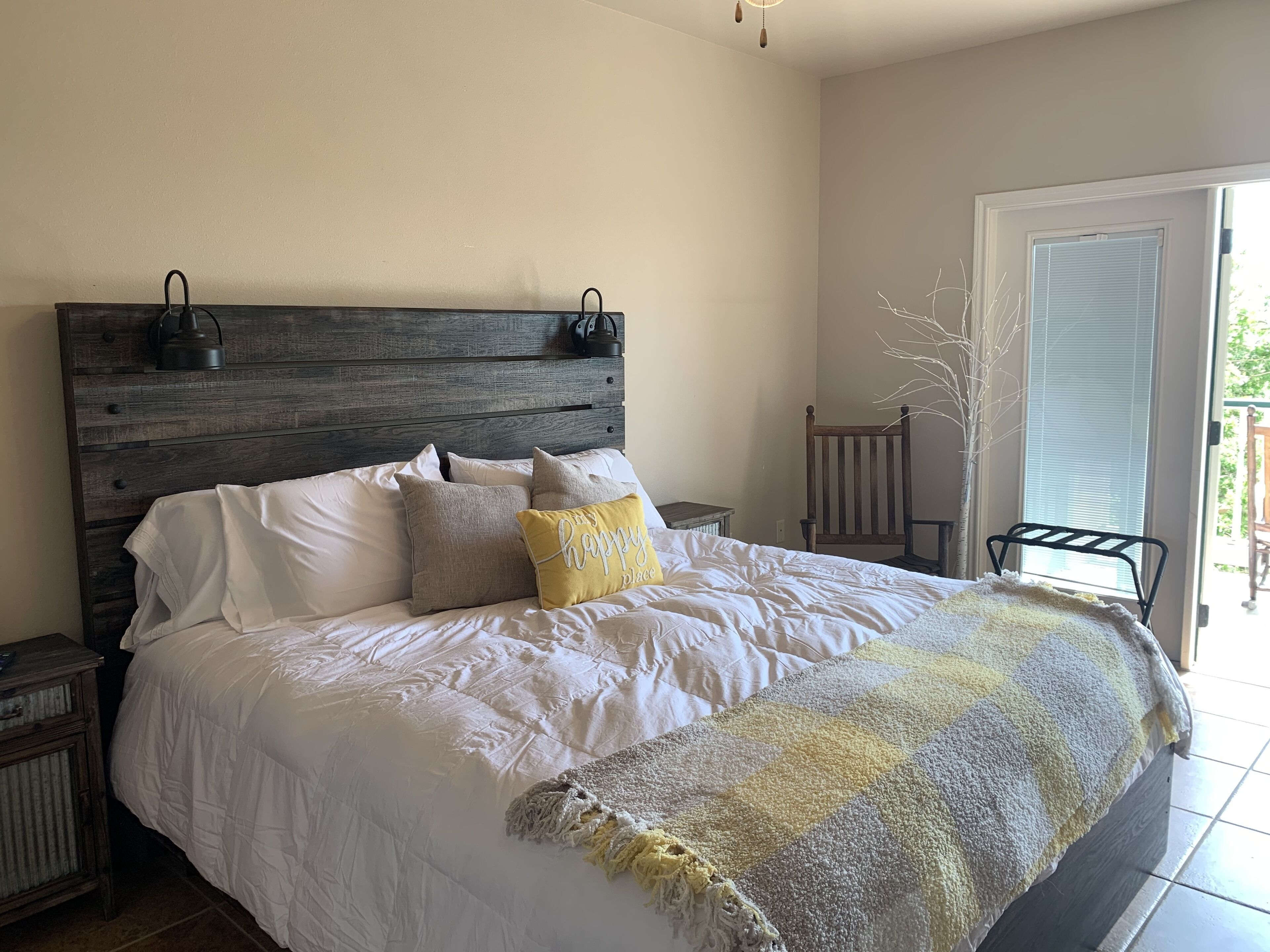 Une chambre à coucher avec une tête de lit en bois, un lit recouvert d’une literie blanche, un oreiller jaune et un plaid à carreaux.