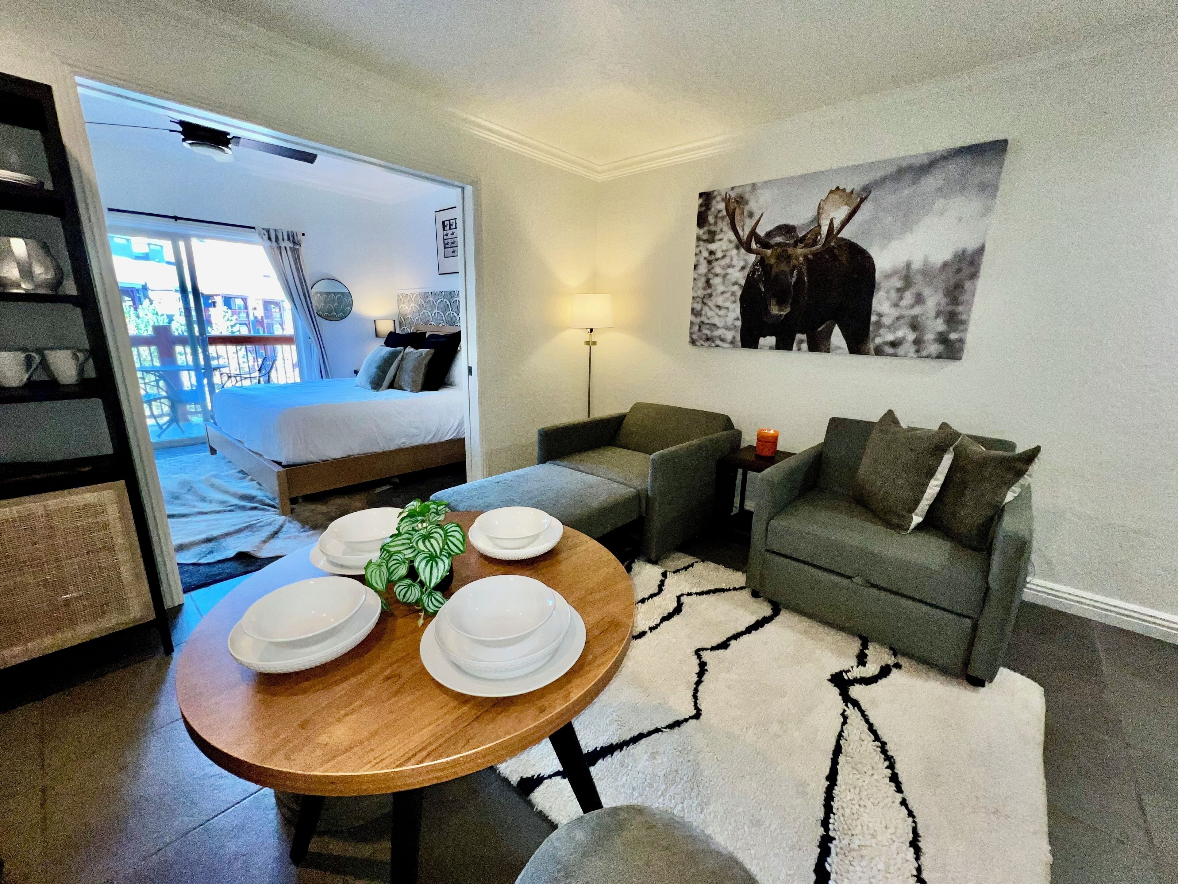 Quarto de hotel moderno com uma cama, um sofá, uma mesa de jantar com pratos, uma planta e uma pintura de um alce.