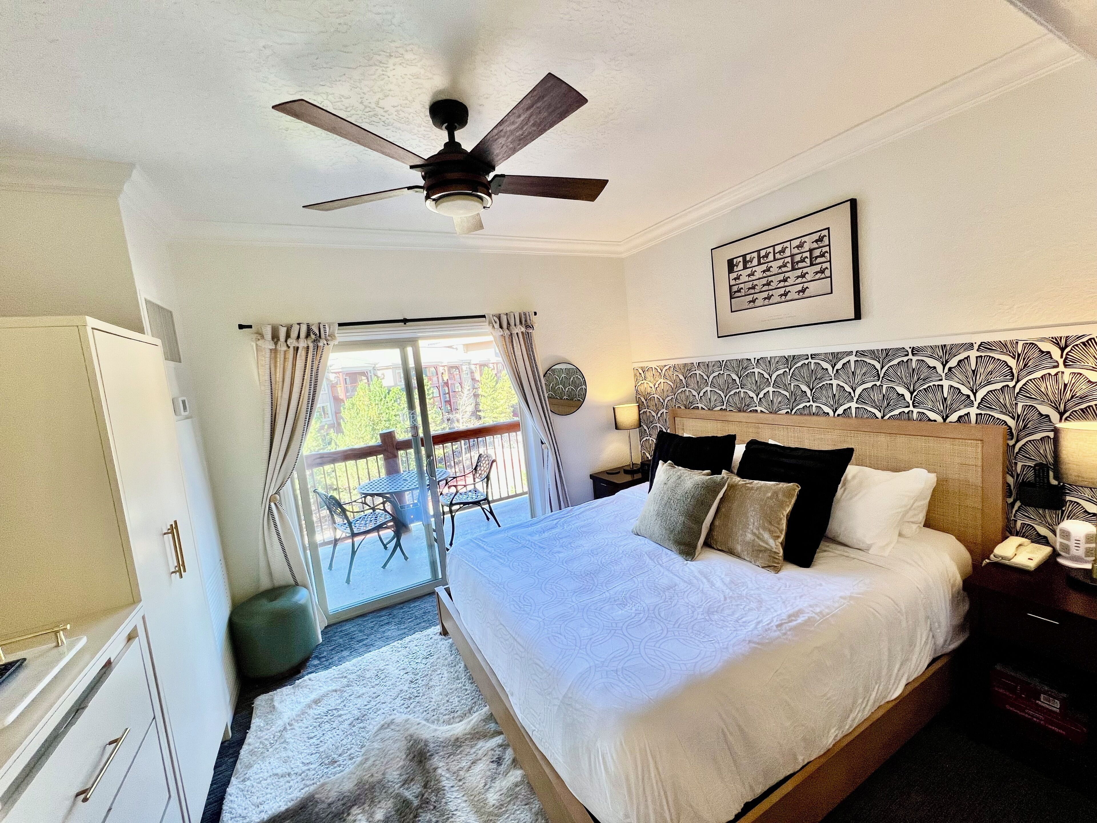Quarto de hotel com cama, ventoinha no teto, varanda com cadeiras e vista para prédios.