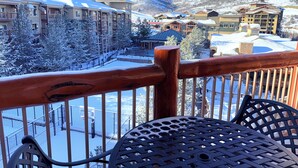 A snow-covered balcony with a view of a ski resort, including buildings and mountains.