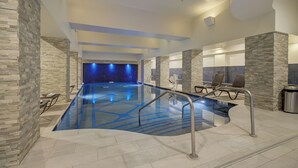 An indoor swimming pool with stone walls, seating by the pool, and a blue-lit backdrop.
