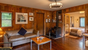 A cozy cabin interior with a leather sofa, a wooden table, a fireplace, and a bed in the adjacent room. - Remote Waterfall Cabin at Swiftwater Acres (Millrift)