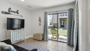 Ein Wohnzimmer mit einem Flachbildfernseher auf einem weißen Schrank, einem Sofa, einem gestreiften Teppich und einer Glasschiebetür, die auf eine Terrasse mit einem Stuhl und einem Korb führt.