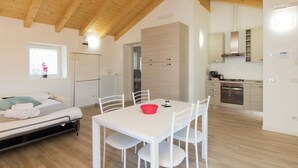 A modern room with a bed, a dining table with chairs, a kitchen area, and a wooden ceiling. - Ciclamino Apartment Residenze Lariane (Colico Piano)