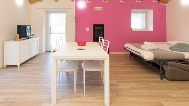 A room with a wooden ceiling, a white table with chairs, a pink wall, a bed, and a TV on a white cabinet. - Ciclamino Apartment Residenze Lariane (Colico Piano)