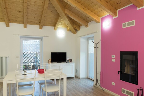 Une pièce avec un plafond en bois, des murs blancs, un mur d’accent rose, un téléviseur posé sur un meuble blanc, une table à manger avec des chaises et une fenêtre dotée d’un moustiquaire métallique.