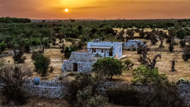 Ein rustikales Steinhaus mit einem modernen Anbau, eingebettet in eine karge, buschige Landschaft unter einem Abendhimmel.