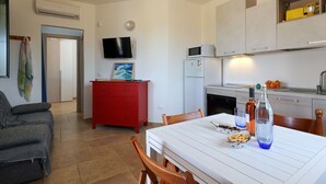 Ein kompakter Wohnbereich mit Küchenzeile, einem Esstisch mit Stühlen, einer roten Kommode, einem Fernseher und einem Bild an der Wand.