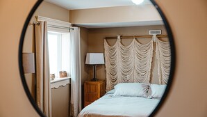 A bedroom with a bed, a nightstand, a lamp, and a macramé headboard. - The Watermark Suite (Drumheller)