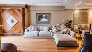 A modern living room with a sectional sofa, wooden accent wall, and a dining area with a table and chairs. - The Watermark Suite (Drumheller)
