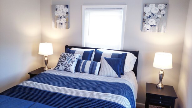 Een net opgemaakt bed met een blauw-witte streepdessin, met aan weerszijden nachtkastjes met lampen, en twee bloemende schilderijen aan de muur.