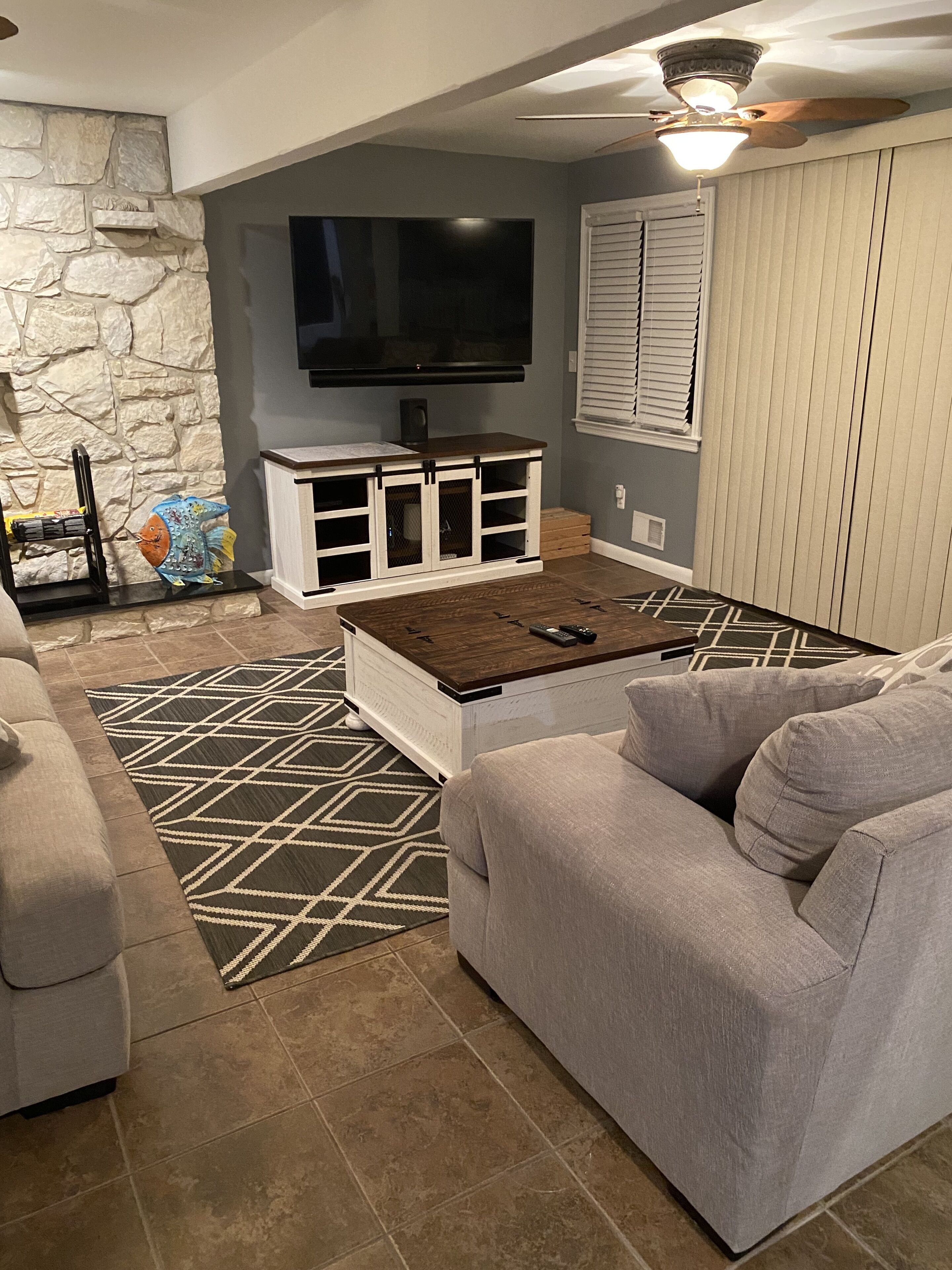 Un salon avec un mur en pierre, un téléviseur à écran plat, un meuble de rangement blanc, un tapis à motifs et un canapé gris.
