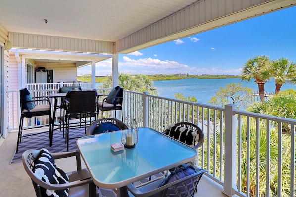 A covered balcony with a table and chairs, overlooking a body of water and greenery.