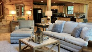 Ein gemütliches Blockhaus-Interieur mit einem Wohnzimmer, das eine Couch, Sessel und einen Couchtisch umfasst. Eine Holztreppe mit Geländern führt auf eine obere Ebene.
