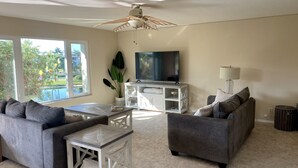 Un salon doté d’un ventilateur de plafond, d’un téléviseur posé sur un meuble blanc, de deux canapés gris, d’une table basse et d’une grande fenêtre donnant sur un paysage verdoyant.