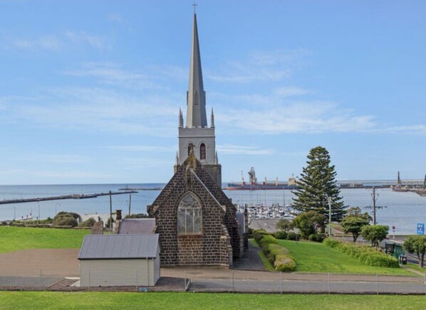 A church with a tall spire, located near a body of water with a bridge and ships in the background.