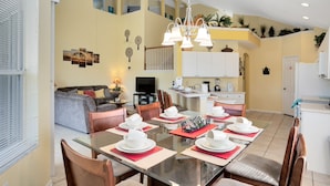 A spacious dining area with a glass table, red placemats, and white dinnerware. A kitchen with white cabinets and a dining table with chairs is visible in the background.