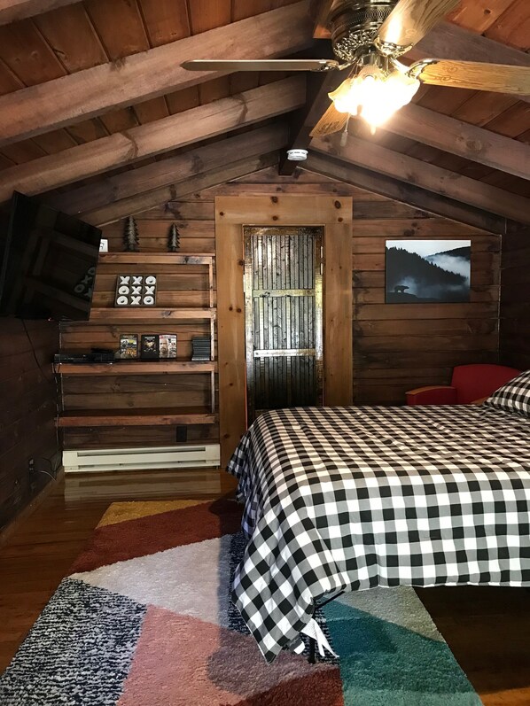 A cozy attic bedroom with a checkered bedspread, a wooden sloped ceiling, a mounted TV, and a framed picture of a bear in a forest.