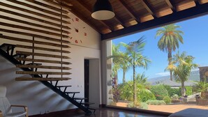 A modern interior with a wooden staircase, a dining area, and a large window offering a view of palm trees and a distant landscape. - Finca La Masía - Relax - Vistas - Internet (La Orotava)