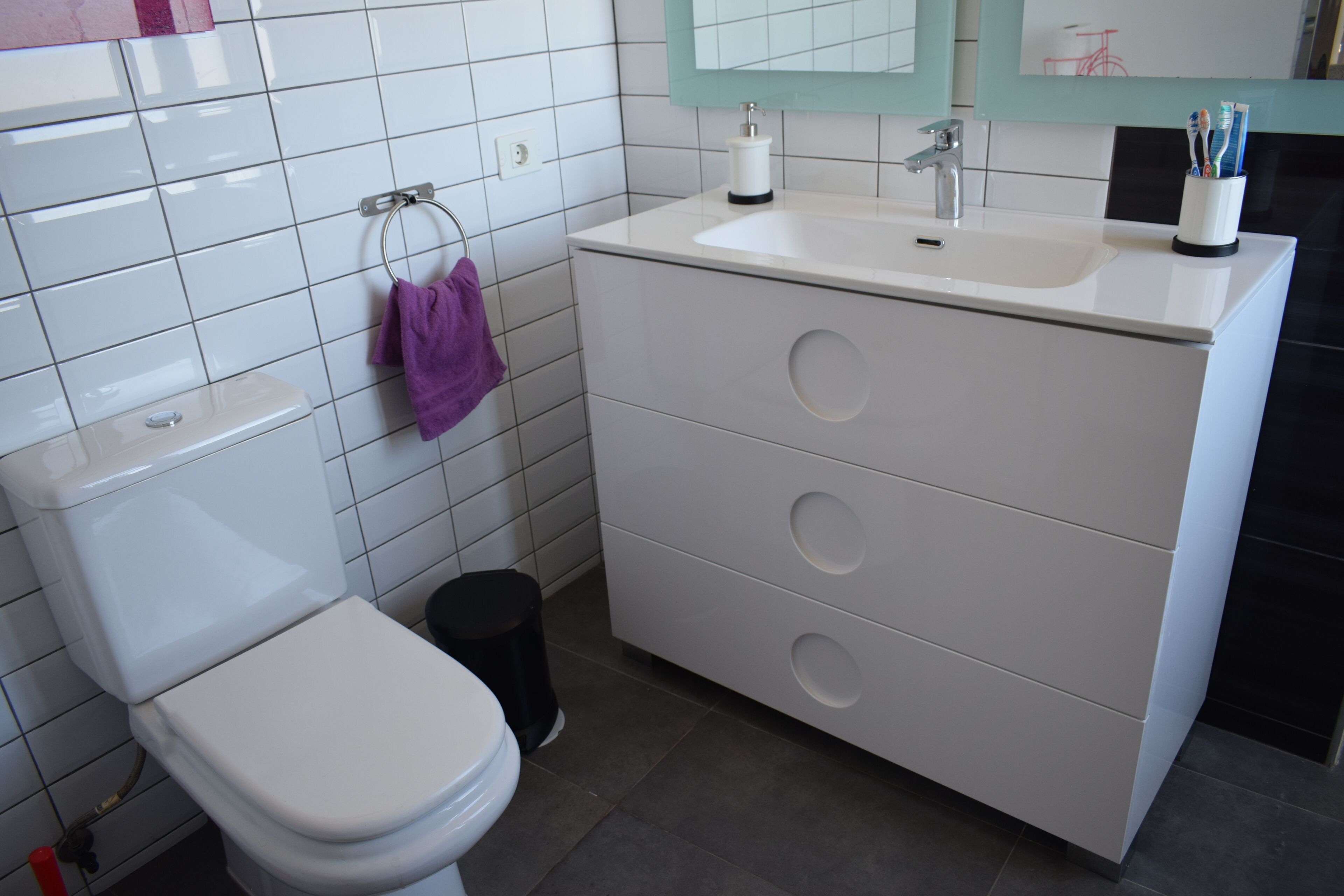 Une salle de bain moderne avec des toilettes blanches, une poubelle noire, un lavabo blanc avec un miroir au-dessus, et un meuble-lavabo avec trois tiroirs.