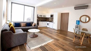 Een moderne woonkamer met een bank, salontafel en een eethoek. Er is ook een keuken met apparatuur en een groot raam.
