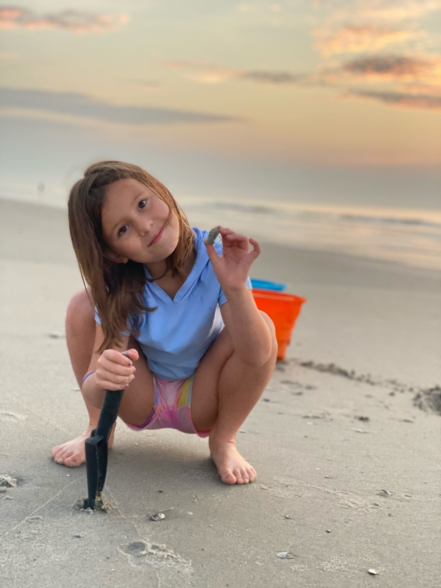 A child squatting on a sandy beach with a colorful bucket in the background.