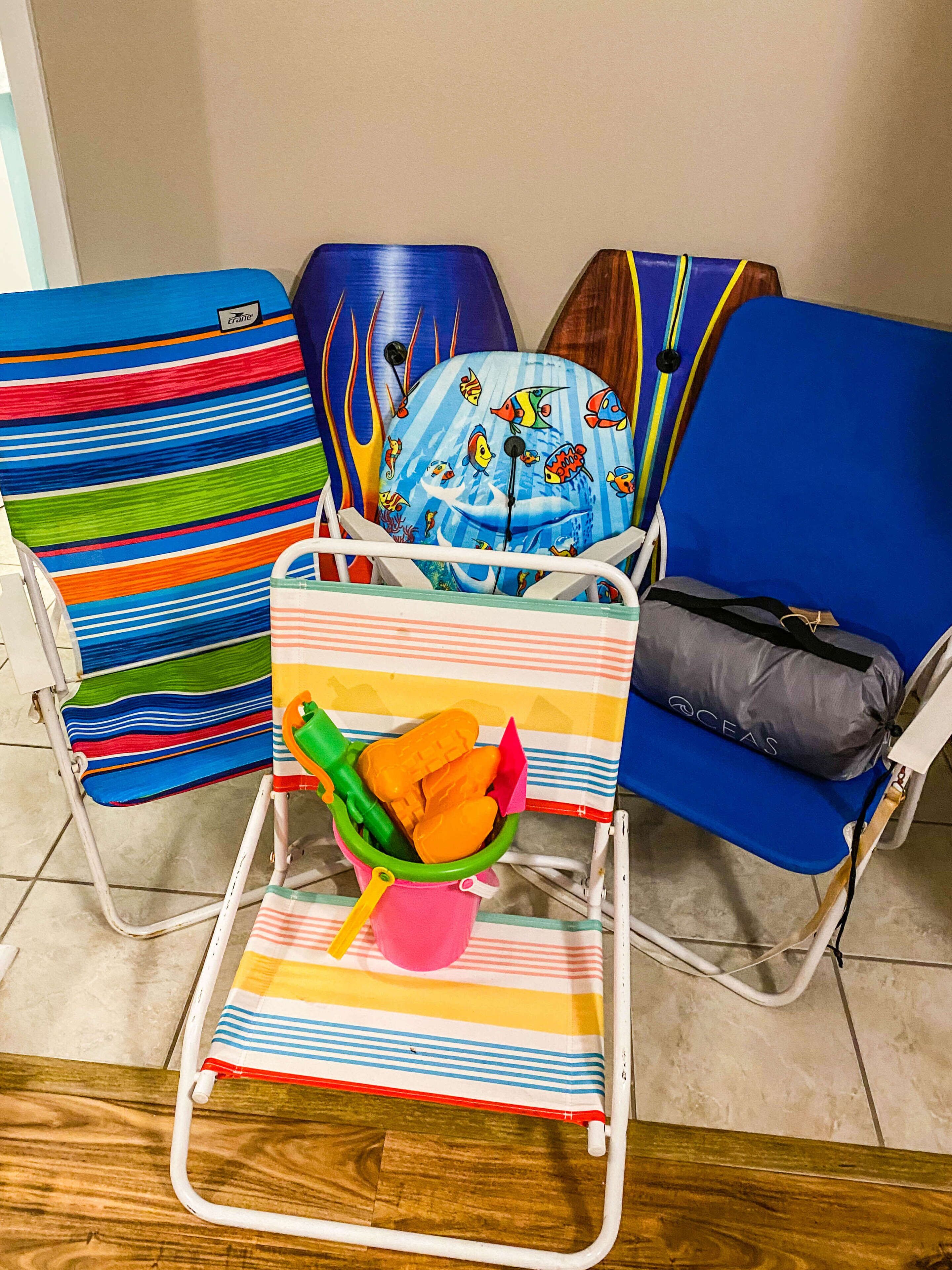 A room with three beach chairs, one with a colorful striped cushion and another with a blue cushion featuring fish and a whale design. A small table with a colorful striped tablecloth and a green bucket containing orange items is in front of the chairs.