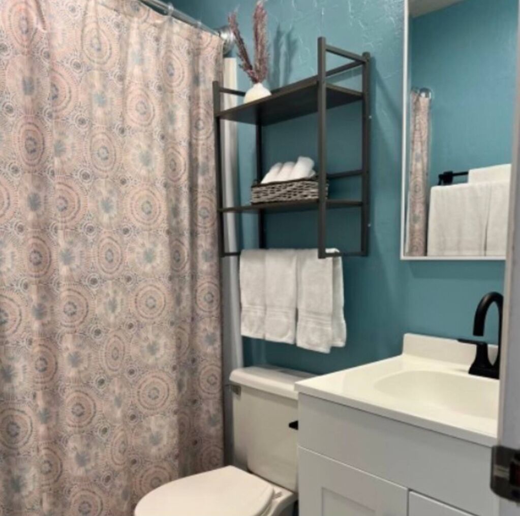 Un baño con cortina de ducha estampada, repisa con toallas, inodoro y lavamanos con espejo.
