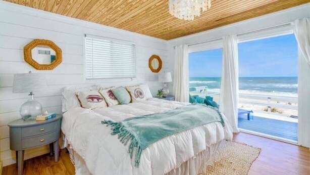 Une chambre avec un plafond en bois, un grand lit recouvert d’une couverture blanche et verte, deux tables de chevet avec des lampes, et une vue sur la plage à travers de grandes fenêtres.
