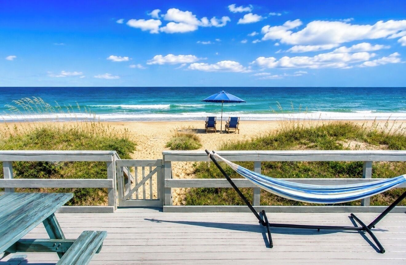Vista de la playa con una hamaca, dos sillas y una sombrilla azul en una terraza de madera.