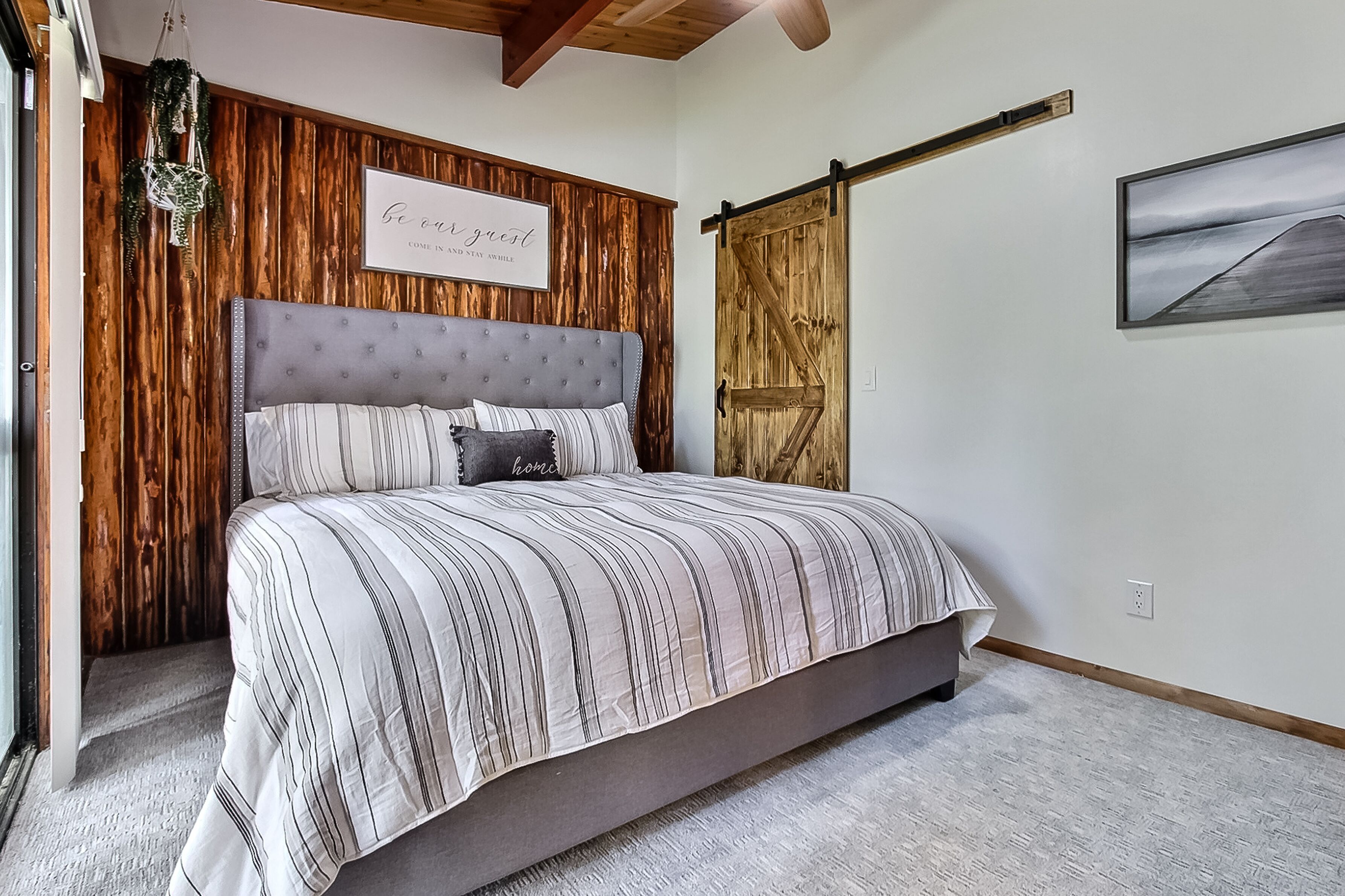 A bedroom with a large bed, wooden panel walls, a 'Come in and stay awhile' sign, a sliding barn door, and a framed picture on the wall.
