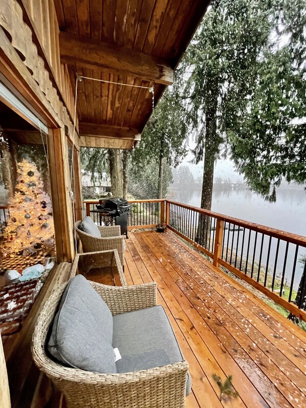 Una terraza de madera con mobiliario de mimbre, una parrilla y vistas a un lago.