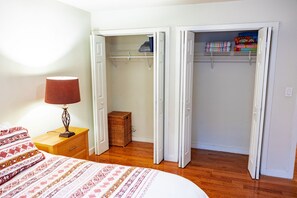 A bedroom with a bed, a nightstand with a lamp, a wooden chest of drawers, a wicker basket, and a closet with open doors revealing folded clothes and shelves. - 2 blocks to the beach- pet friendly with parking! (Asbury Park)