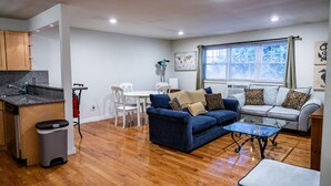 Una sala moderna con un sofá azul, una mesa de centro de vidrio y un sofá blanco. Hay una mesa de comedor con sillas, una zona de cocina con armarios y una ventana con cortinas.