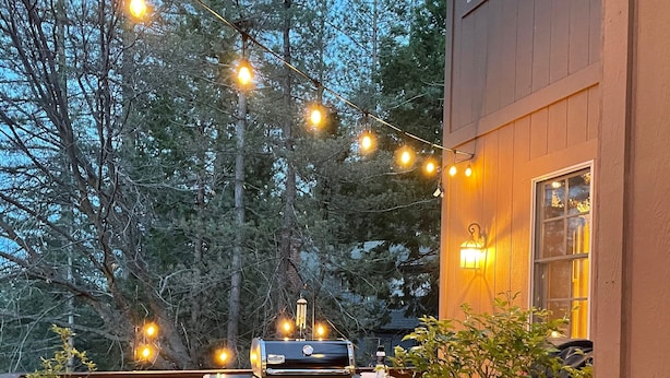 Une terrasse en bois avec un barbecue et des guirlandes lumineuses, entourée d’arbres.