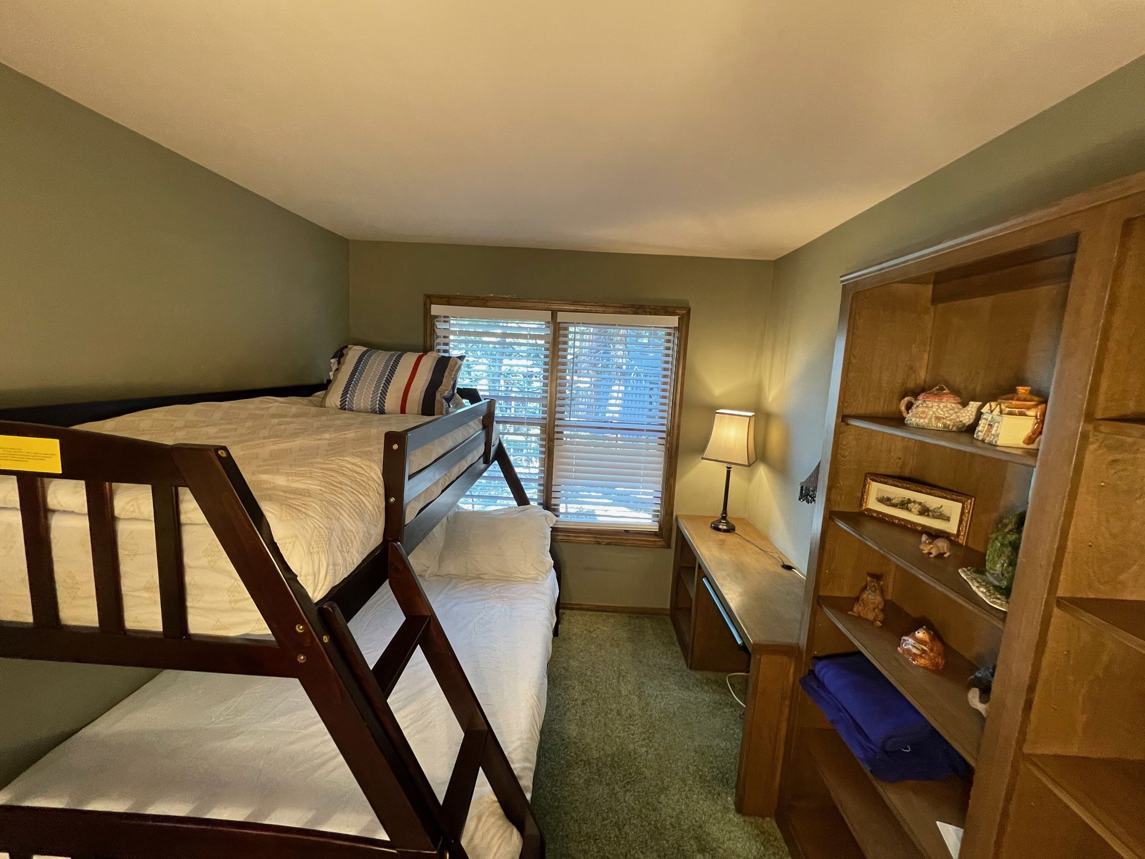 A bedroom with a bunk bed, a bedside table with a lamp, a wooden wardrobe, and a window with blinds.
