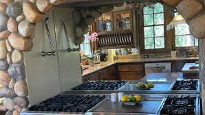 Une cuisine avec un mur en pierre, plusieurs cuisinières à gaz, des armoires en bois et une fenêtre donnant sur des arbres.