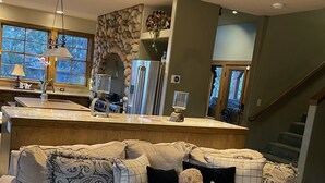 Un salon avec un canapé d’angle beige, un coin cuisine avec un mur en pierre et un escalier menant à un autre niveau.