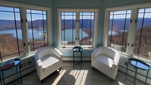 Une pièce comprenant trois fauteuils blancs, une petite table et de grandes fenêtres donnant sur un lac et des montagnes.