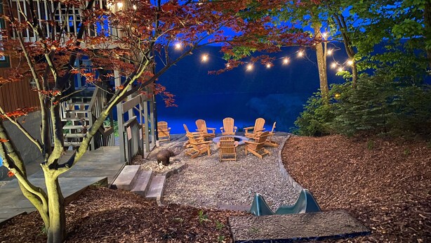 Un espace extérieur aménagé avec des chaises Adirondack et un foyer, entouré d’arbres et illuminé de guirlandes lumineuses.